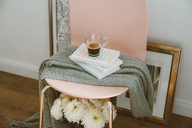 A serene self-care corner with a cozy gray blanket over a pink chair, a book on therapy, and a cup of coffee, next to a bouquet of white chrysanthemums, symbolizing personal time for reflection and healing after the loss of a parent.