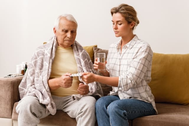 A compassionate caregiver offering medication to an elderly man wrapped in a blanket, depicting a scene of anticipatory grief and the challenges of caregiver burnout.