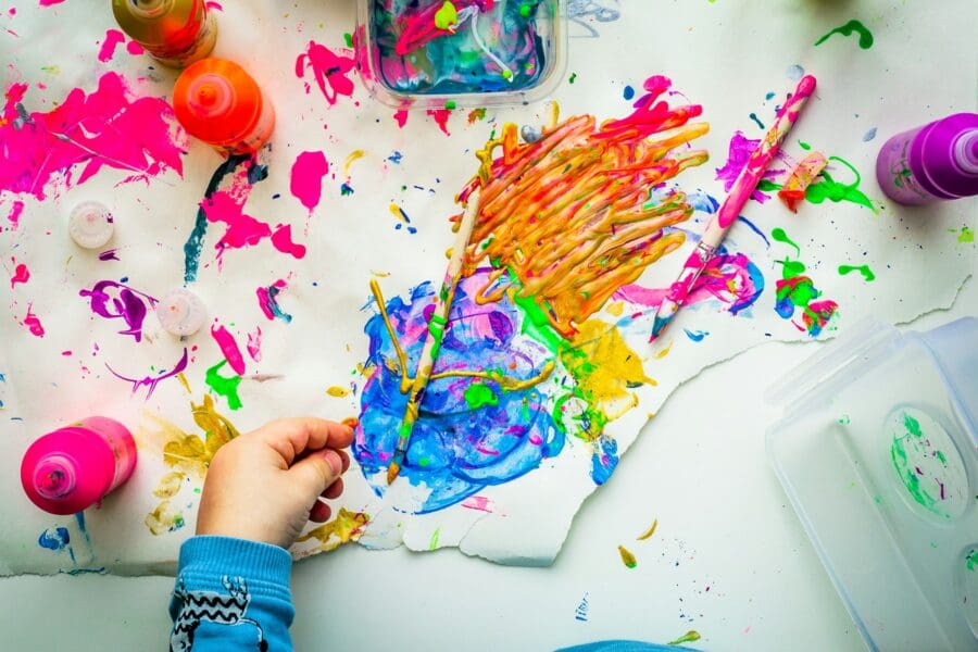 A child's hand creating a colorful painting, surrounded by vibrant splashes of paint and art supplies, depicting the use of art as a form of expression and healing for grieving children.