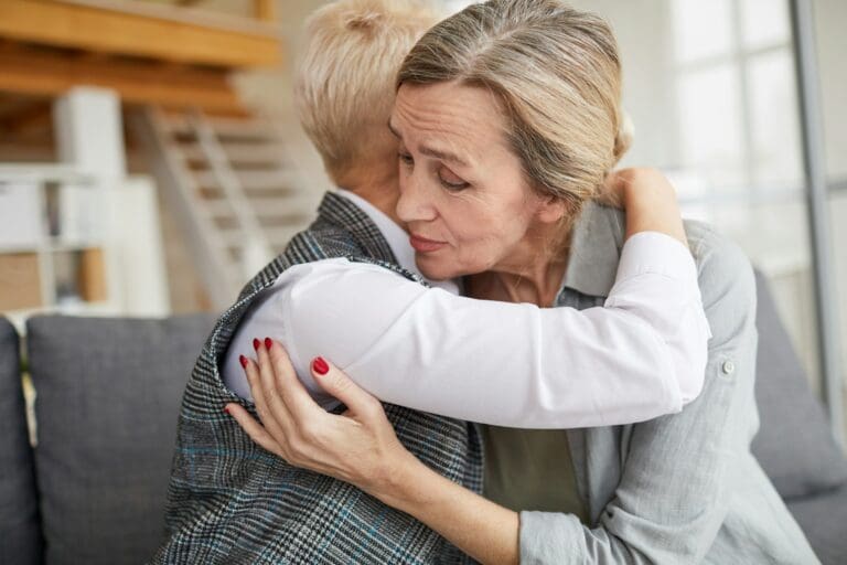 Two women embracing in a comforting hug, one with a pensive expression, embodying the emotional support necessary in understanding that acceptance of loss doesn't equate to 'getting over' grief.