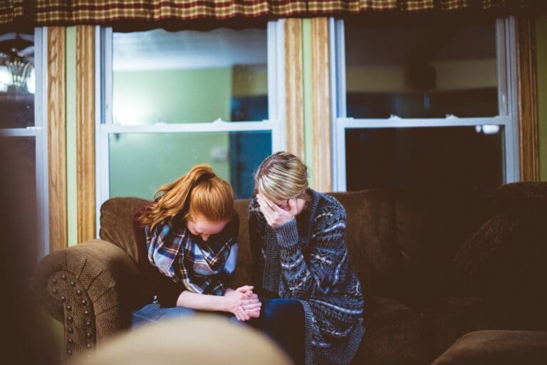 Two people sitting close on a sofa, holding hands, one with their head bowed and the other with a hand covering their face, in a moment that suggests the emotional strain of parent-child estrangement.