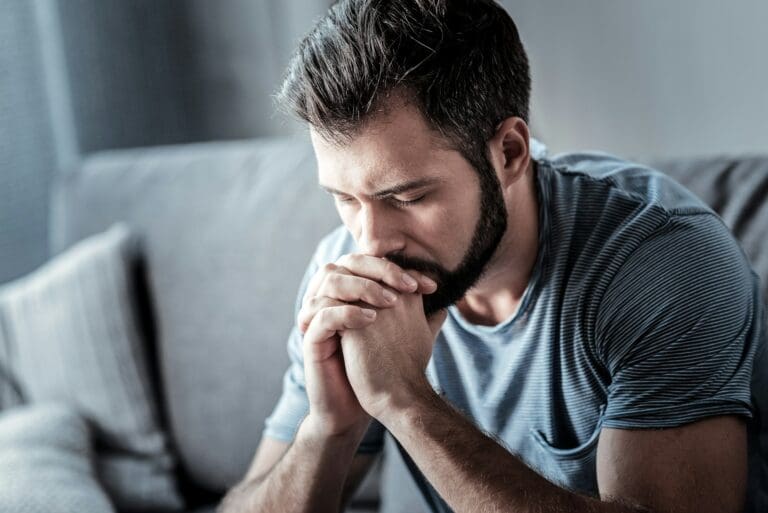 A distressed man sitting on a couch, hands clasped in prayer, deeply contemplating the complex emotions of grieving an abuser, illustrating the solitude and confusion that can accompany such a loss.