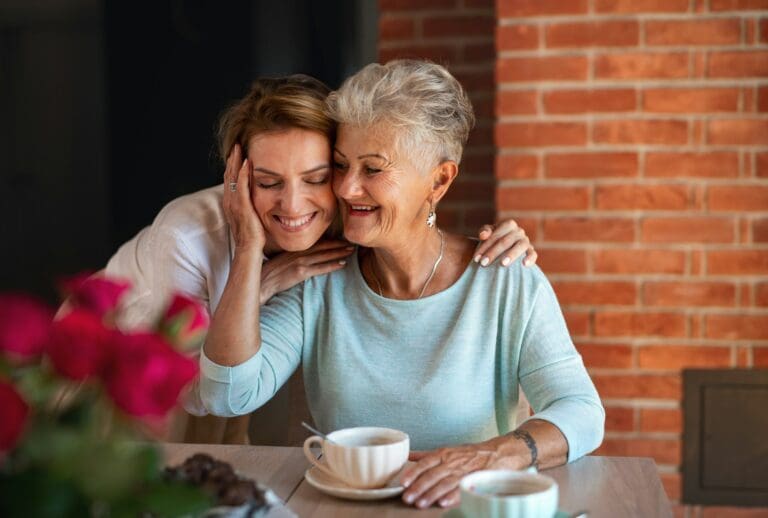 A young woman affectionately resting her head on her elderly mother's shoulder as they share a moment of connection over coffee, representing support and understanding in times of grief.